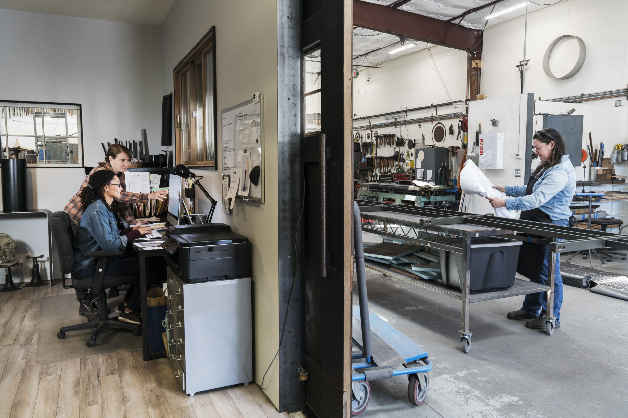 Metal Workshop,USA,Two women at a desk in office area of a metal workshop, woman standing at mobile workbench.